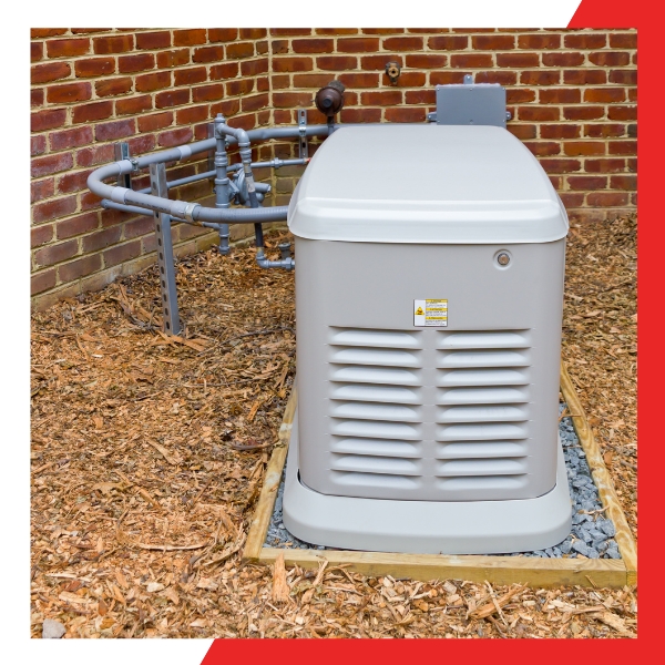 A Generac home standby generator, light beige with a gray top, sits on a gravel base next to a brick house with exposed piping, surrounded by wood chips.