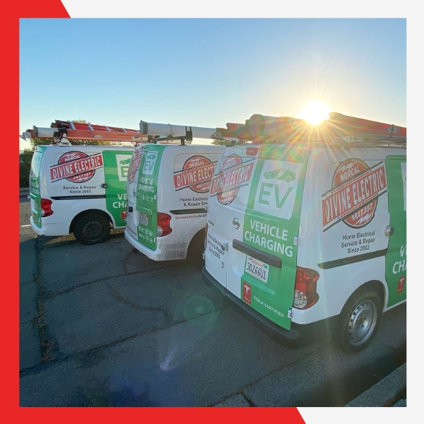 Three white service vans with green and white "Divine Electric" logos and "Vehicle Charging" text, parked side-by-side with ladders on top, illuminated by a bright sunflare.