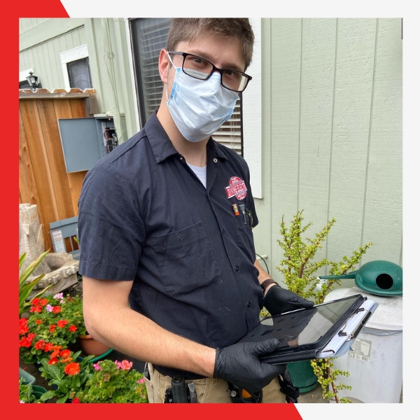 A male technician wearing glasses, a face mask, and gloves, holding a tablet and clipboard, standing outside a house next to some plants and outdoor equipment.