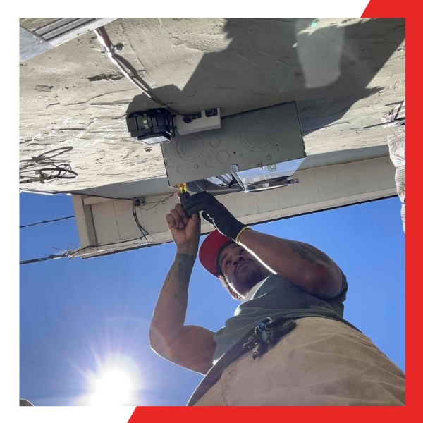 A low-angle shot of a technician, wearing gloves and a red cap, working on an electrical panel mounted on the underside of an outdoor eaves or structure, with the sun brightly shining behind them.