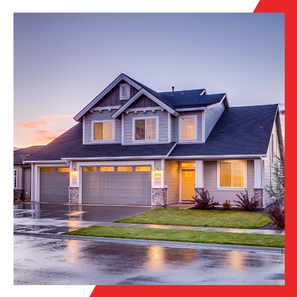 A modern two-story suburban house with a gray exterior and dark roof, illuminated windows at dusk, and a wet driveway reflecting the twilight sky.