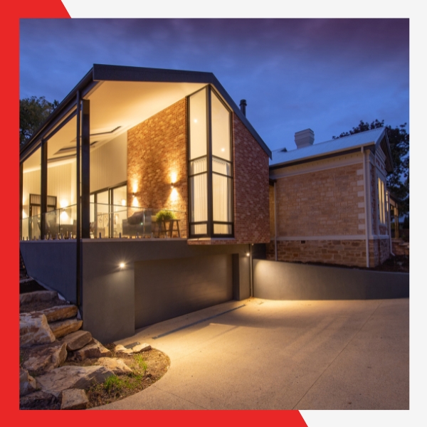 A modern, illuminated house at dusk with large windows, brick and dark paneling, and an inviting driveway lit by ground lights.
