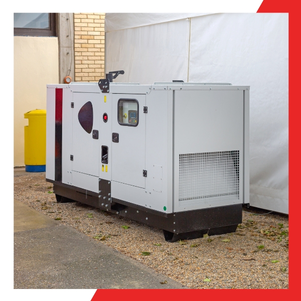 A large, industrial-looking standby generator, light gray with some red and yellow markings, sitting on a gravel patch outside a building with a yellow bin and white tarp in the background.