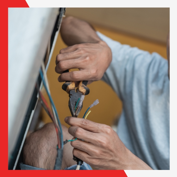 Close-up of a person's hands wearing a t-shirt, using pliers to work on colored electrical wires connected to a utility box or appliance.